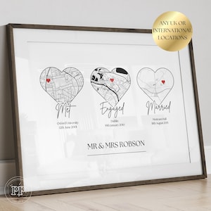 May include: A framed print featuring three heart-shaped map illustrations, each with a red heart marking a location. Text includes "Met," "Engaged," and "Married," along with dates and locations. The print is framed in a dark brown wooden frame.