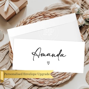 May include: White envelope and card with the name "Amanda" in elegant black script and a small heart. The card is part of a personalized envelope upgrade, resting on a woven placemat with a gift box and flowers in the background.