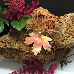 May include: A gold chain necklace with a large, copper-colored maple leaf pendant. The leaf has a textured, iridescent finish and is set against a background of brown rock and pink flowers.