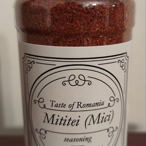 May include: A glass jar with a red lid filled with brown seasoning. The label reads "Taste of Romania Mititei (Mici) seasoning Net Wt. 4 Oz (113g)"