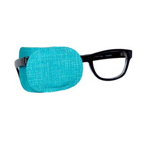May include: Black framed spectacles with a turquoise eye patch covering the left lens. The eye patch is made of a soft, textured fabric.