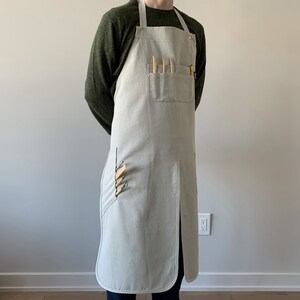 May include: A light gray canvas apron with a large pocket and multiple tool slots. The apron has a long strap that ties around the neck and waist.