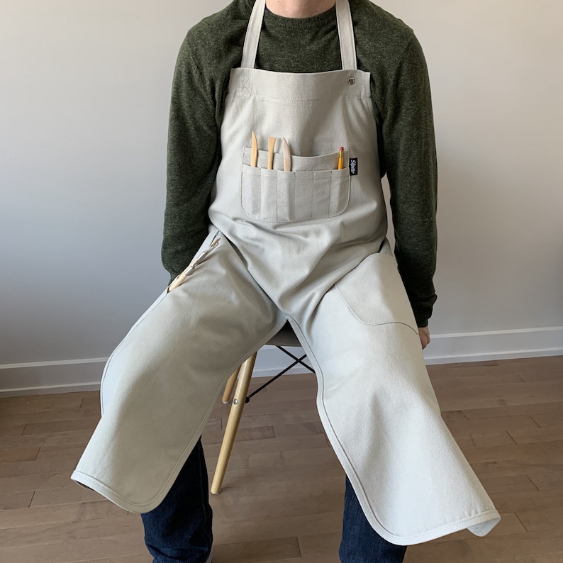 May include: A light gray artist's apron with multiple pockets and a tool holder. The apron is worn by a person with a green shirt.