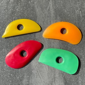 Puede incluir: Cuatro herramientas de costilla de cerámica de colores amarillo, naranja, rojo y verde. Cada herramienta tiene una forma curva con un agujero central. Estas herramientas se utilizan para dar forma y alisar la arcilla en cerámica.