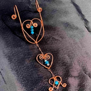 May include: Copper wire art piece featuring three heart shapes and decorative swirls, strung together with a chain. The hearts are adorned with turquoise-colored beads. The piece is displayed against a dark background.