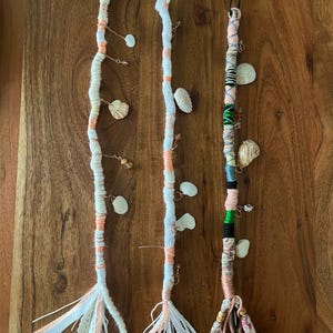 May include: Three white and pink hair wraps with shells and beads. The wraps are tied with a bow at the bottom.