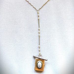 May include: A gold-toned locket necklace with a pearl accent. The necklace features a delicate chain with small pearls and a rectangular locket pendant with an oval pearl center. The locket has a silver bar across the top.