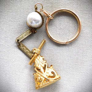 Puede incluir: Un llavero dorado con una perla grande y redonda y un dije decorativo y ornamentado. El llavero incluye un anillo dorado y un dije detallado de estilo vintage con diseños intrincados. La perla es blanca y el metal tiene un acabado pulido.