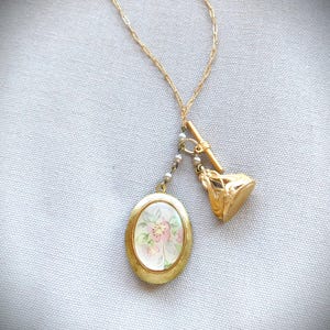 May include: A gold-toned necklace featuring a floral locket pendant and a decorative charm. The oval locket displays a pink and white floral design. The necklace has a delicate chain and a small pearl accent.