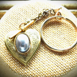 May include: A gold-colored heart-shaped locket with a silver-colored oval stone in the center. The locket is attached to a chain and a gold-colored key ring. The locket has an engraved design and is resting on a tan woven surface.