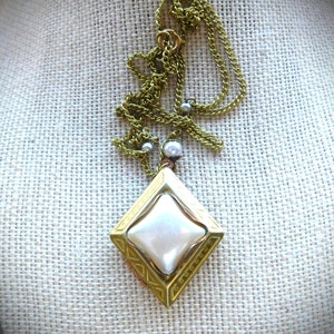 Puede incluir: Un collar de cadena dorada con un colgante en forma de diamante que presenta una perla blanca en el centro. El colgante tiene un borde decorativo.