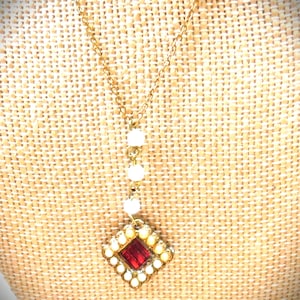 May include: A gold-toned necklace with a pendant featuring a red rectangular stone surrounded by small white beads. The pendant is diamond-shaped. The necklace has a delicate chain and a vertical strand of white beads.