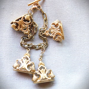 May include: Gold-toned antique cufflinks with ornate filigree detailing. The cufflinks feature a chain and a T-bar closure. The design includes floral and scrollwork elements, creating an elegant and vintage aesthetic.