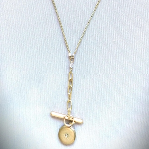 May include: A gold-toned lariat necklace featuring a delicate chain, a small clear stone accent, and a toggle clasp closure. A circular gold-toned pendant hangs from the toggle bar, centered with a single small, sparkling clear stone.