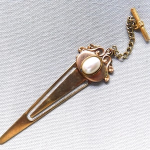May include: Vintage gold-toned tie clip with a decorative oval pearl center and a chain. The clip has an ornate design around the pearl and a T-bar closure. The clip is approximately 3 inches long.