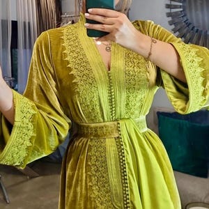 A woman wearing a chartreuse velvet caftan with wide sleeves and detailed embroidery on the neckline, bodice, and belt, taking a mirror selfie.