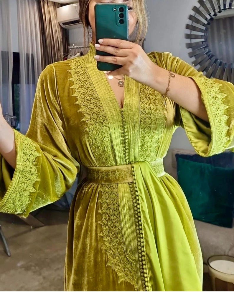 A woman wearing a chartreuse velvet caftan with wide sleeves and detailed embroidery on the neckline, bodice, and belt, taking a mirror selfie.