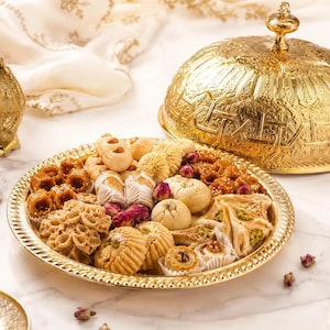 Peut inclure: Un plateau de service doré orné rempli d'un assortiment de pâtisseries. Le plateau rond a un bord décoratif et est accompagné d'un couvercle doré assorti. Les pâtisseries sont de formes et de couleurs variées, certaines décorées de noix et de graines.