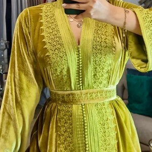 A woman in a chartreuse velvet caftan with a V-neck and embroidered details on the bodice, sleeves, and belt, taking a mirror selfie.