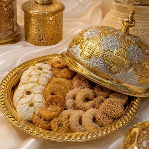 Peut inclure: Un plateau de service rond en métal doré avec un couvercle bombé assorti présentant des motifs géométriques argentés et dorés complexes. Le plateau est rempli d'un assortiment de biscuits ronds et en forme de croissant, certains garnis de noix.