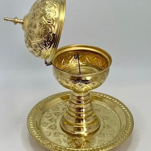 Op de afbeelding: Een gouden metalen wierookbrander met een koepelvormig deksel en een decoratief dienblad. De brander heeft ingewikkelde gegraveerde ontwerpen en een dunne draad erin.
