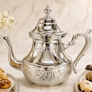 May include: A shiny silver teapot with ornate detailing, including a decorative handle and spout. The teapot is placed on a white surface, with a blurred background. The image suggests a theme of tea service or kitchenware.