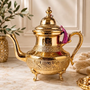 May include: A gold-colored teapot with intricate floral and geometric patterns. The teapot has a curved spout, a decorative handle, and a lid with a small finial. A purple tassel hangs from the lid. The teapot sits on three small feet.