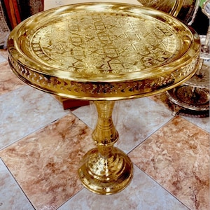 Può includere: Un tavolo in metallo dorato con un design intricato e decorato sulla parte superiore. Il tavolo ha un piano rotondo e un unico piedistallo decorativo.