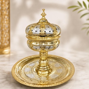 May include: A gold and silver metal incense burner with a matching circular tray. The burner features a removable lid with a pointed finial, intricate floral engravings, and small circular cutouts, all supported by a pedestal base.