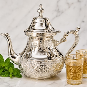 Peut inclure: Une théière argentée avec des motifs floraux et géométriques complexes, un bec verseur incurvé et une poignée décorative. Deux petits verres avec des motifs de filigrane dorés sont à côté de la théière. Des feuilles de menthe fraîche sont à gauche. Design marocain traditionnel.