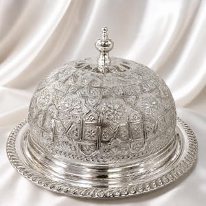 May include: A silver-colored, domed serving dish with intricate embossed floral and geometric patterns. The dish includes a round base with a decorative rope-like edge and a matching lid with a small, ornate handle. The background is a white, silky fabric.