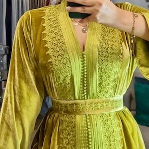 A woman wearing a chartreuse velvet caftan with intricate embroidery on the V-neckline, sleeves, and belt, holding a dark green phone to take a mirror selfie.