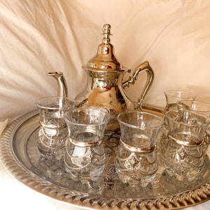 Pode incluir: Um conjunto de chá marroquino de prata com uma chaleira e seis copos em uma bandeja de prata. A chaleira e os copos têm desenhos florais intrincados.