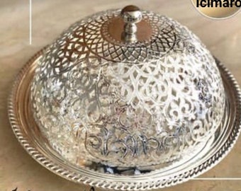 Può includere: Un piatto da portata argentato e ornato con un coperchio a cupola. Il coperchio e la base presentano intricati motivi traforati. Il piatto poggia su un vassoio rotondo argentato con un bordo decorativo a forma di corda. Un piccolo pomello rotondo sovrasta il coperchio.