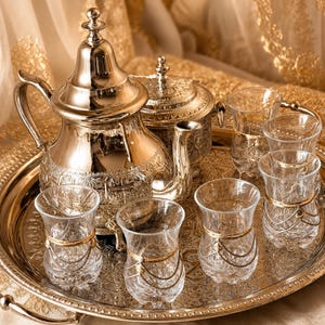 May include: A silver tea set on a silver tray. The set includes a teapot, sugar bowl, and six small glasses with gold accents. The background is a gold-coloured fabric.