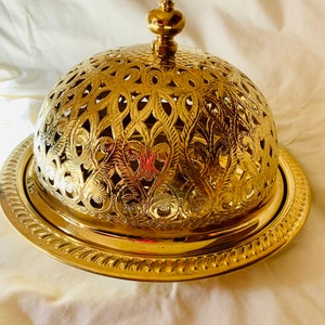 Puede incluir: Un plato de servir de metal dorado con una tapa abovedada. La tapa tiene un diseño intrincado, decorativo y recortado. El plato tiene un borde elevado.