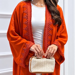 Peut inclure: Une abaya orange vif ouverte sur le devant, ornée de broderies orange foncé complexes le long des revers et des poignets, portée avec un haut blanc et un petit sac à main structuré en paille beige avec des finitions en cuir marron.
