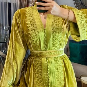 A woman in a chartreuse velvet caftan with embroidered V-neck, sleeves, and a matching belt, holding a dark green phone for a mirror selfie.