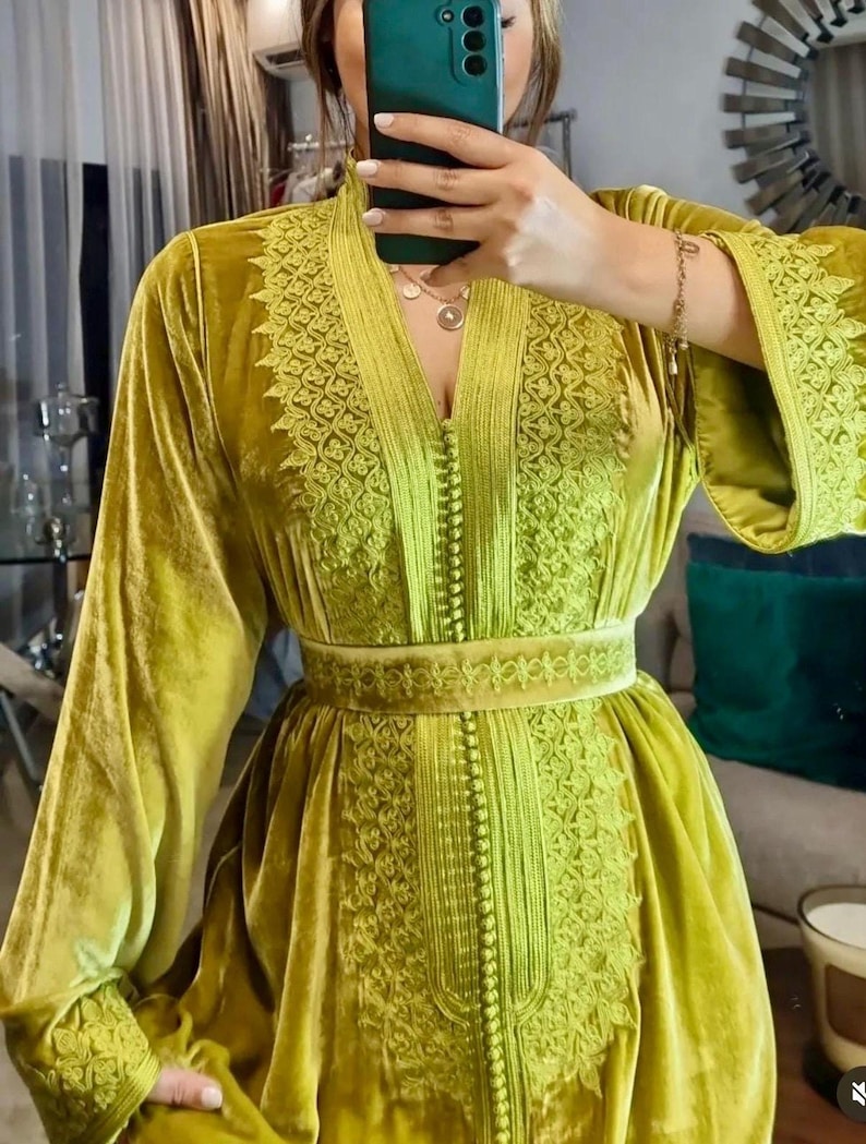 A woman in a chartreuse velvet caftan with embroidered V-neck, sleeves, and a matching belt, holding a dark green phone for a mirror selfie.