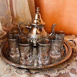 May include: A silver tea set with a teapot, six glasses, and a serving tray. The teapot and tray have ornate designs. The glasses have a decorative pattern. The set is displayed on a patterned surface.