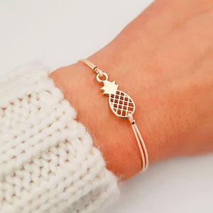May include: A gold pineapple charm bracelet with a beige cord. The bracelet is adjustable and has a sliding knot.