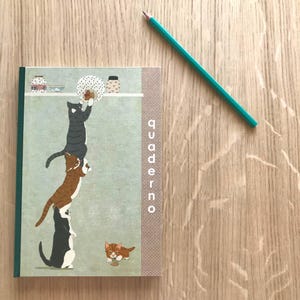 May include: A notebook with a green spine and a cover featuring a whimsical illustration of cats stacked on top of each other. The word "quaderno" is printed vertically on the side. A teal pencil rests on a wooden surface.