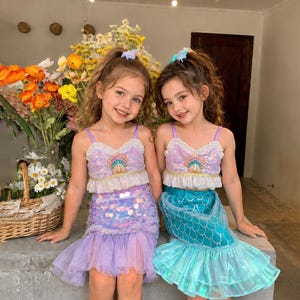 Könnte beinhalten: Zwei passende zweiteilige Outfits mit Pailletten-Bauchfrei-Tops mit Rüschenbesatz und muschelförmigen Verzierungen. Ein Outfit umfasst einen lavendelfarbenen Paillettenrock mit Tüllsaum, das andere einen türkisfarbenen Paillettenrock mit Tüllsaum.