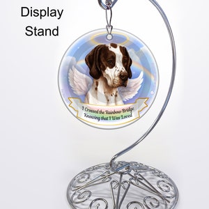 May include: A round, white, glass ornament with a brown and white dog with angel wings. The dog is looking to the right. The ornament has a rainbow and clouds in the background. The text "I crossed the Rainbow Bridge Knowing that I was Loved" is on a banner below the dog. The ornament is on a silver metal stand.