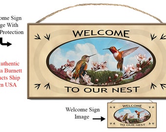 Puede incluir: Un letrero de bienvenida con fondo beige y marco marrón. El letrero presenta una pintura de dos colibríes posados en una rama con flores rosadas. El texto "WELCOME TO OUR NEST" está impreso en letras negras debajo de la pintura.