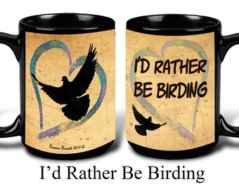 Puede incluir: Taza de cerámica negra con el texto "I'd Rather Be Birding". La taza presenta la silueta de un pájaro dentro de un diseño en forma de corazón con detalles en azul y morado. La taza tiene un asa negra.