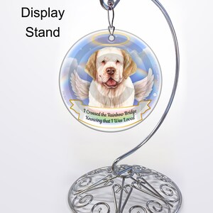 May include: A round ornament with a dog angel design, displayed on a silver metal stand. The ornament has the text "I Crossed the Rainbow Bridge Knowing that I Was Loved". The stand is a decorative display for ornaments.