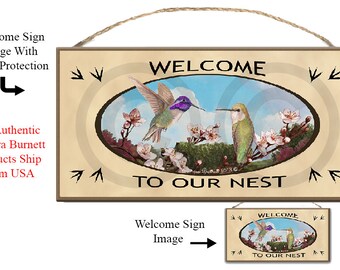 Puede incluir: Un letrero de bienvenida de madera con fondo beige que presenta dos colibríes en vuelo. Los colibríes están posados en una rama con flores rosas. El letrero dice "Bienvenido a nuestro nido" en una fuente marrón.