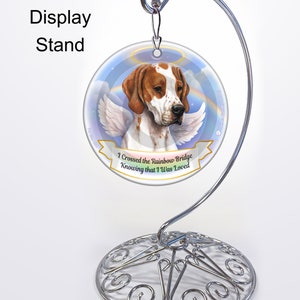 May include: A white and brown dog with angel wings on a round ornament with a blue sky and rainbow background. The ornament has the text "I crossed the Rainbow Bridge Knowing that I was Loved". The ornament is on a silver metal stand.