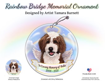 Op de afbeelding: Een ronde, glazen ornament met een regenboogbrug en wolken. Een bruine en witte hond met engelenvleugels bevindt zich in het midden. De tekst "In Loving Memory of Bella 2006-2021" staat op de ornament.